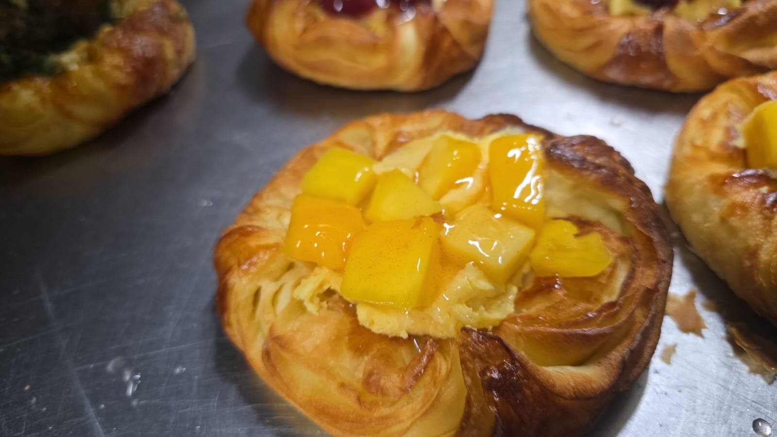 A mango danish topped with diced mango and vanilla custard on a steel bench