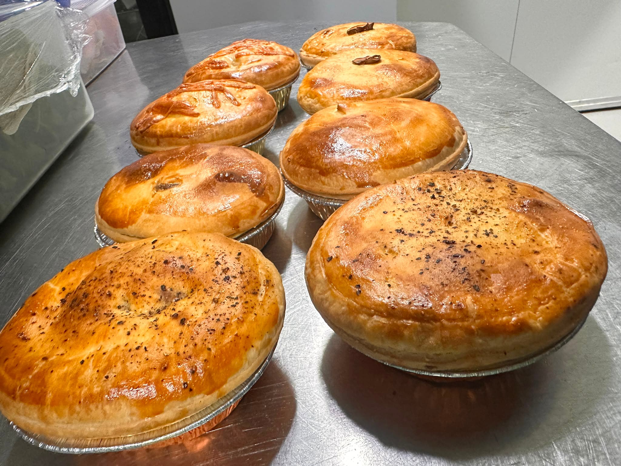 A row of pies in foil trays — pepper, chunky beef and chicken, glazed and cracked with black pepper