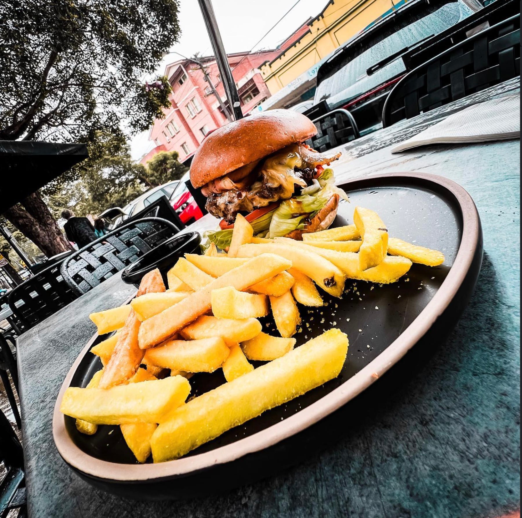 A burger and thick-cut chips on a black plate at an outdoor table on Burlington Road