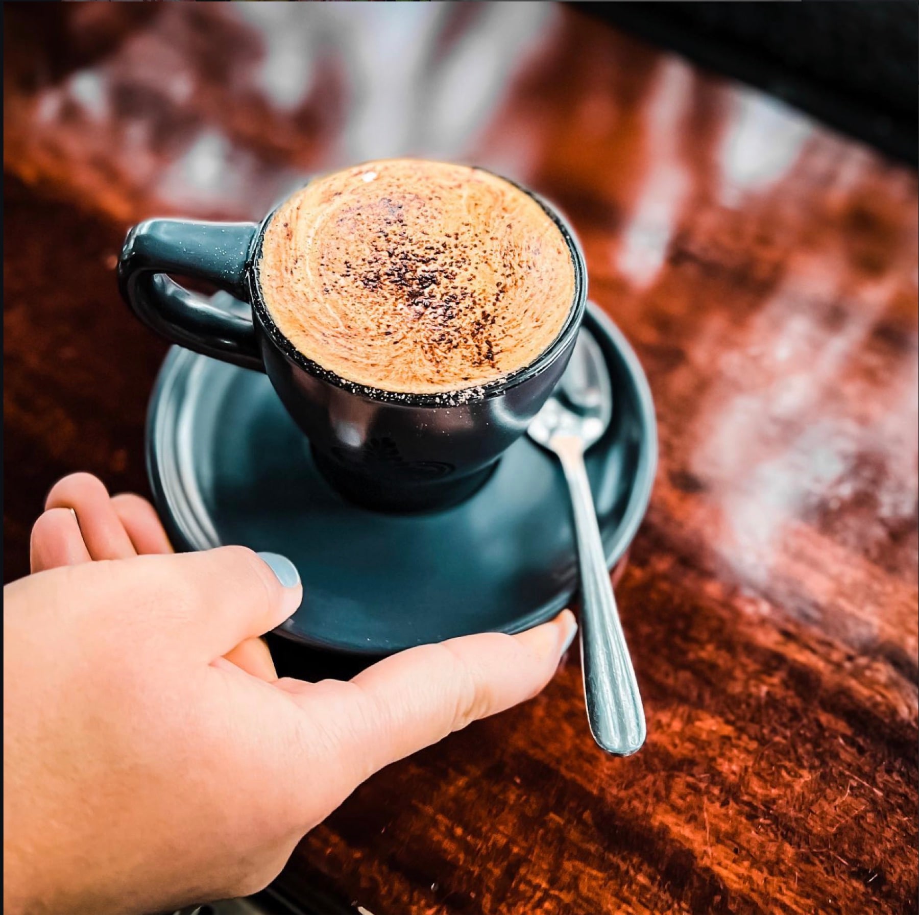 Two hands resting either side of a cappuccino on a dark saucer, on a polished timber counter