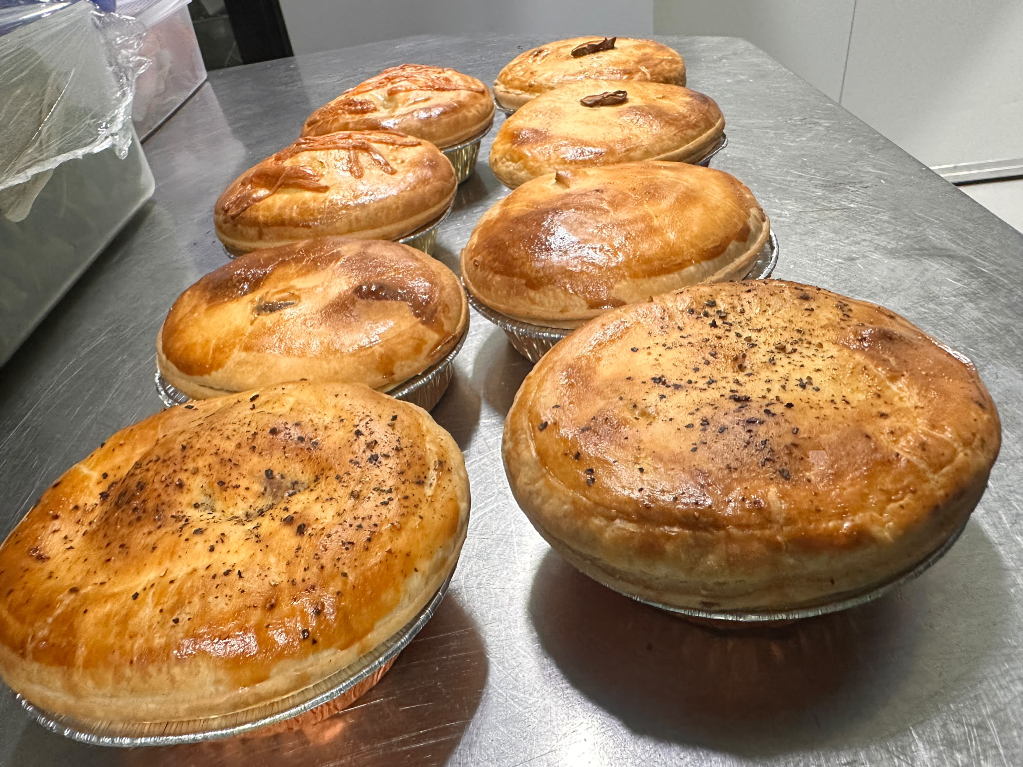 A row of pies in foil trays — pepper, chunky beef and chicken, glazed and cracked with black pepper