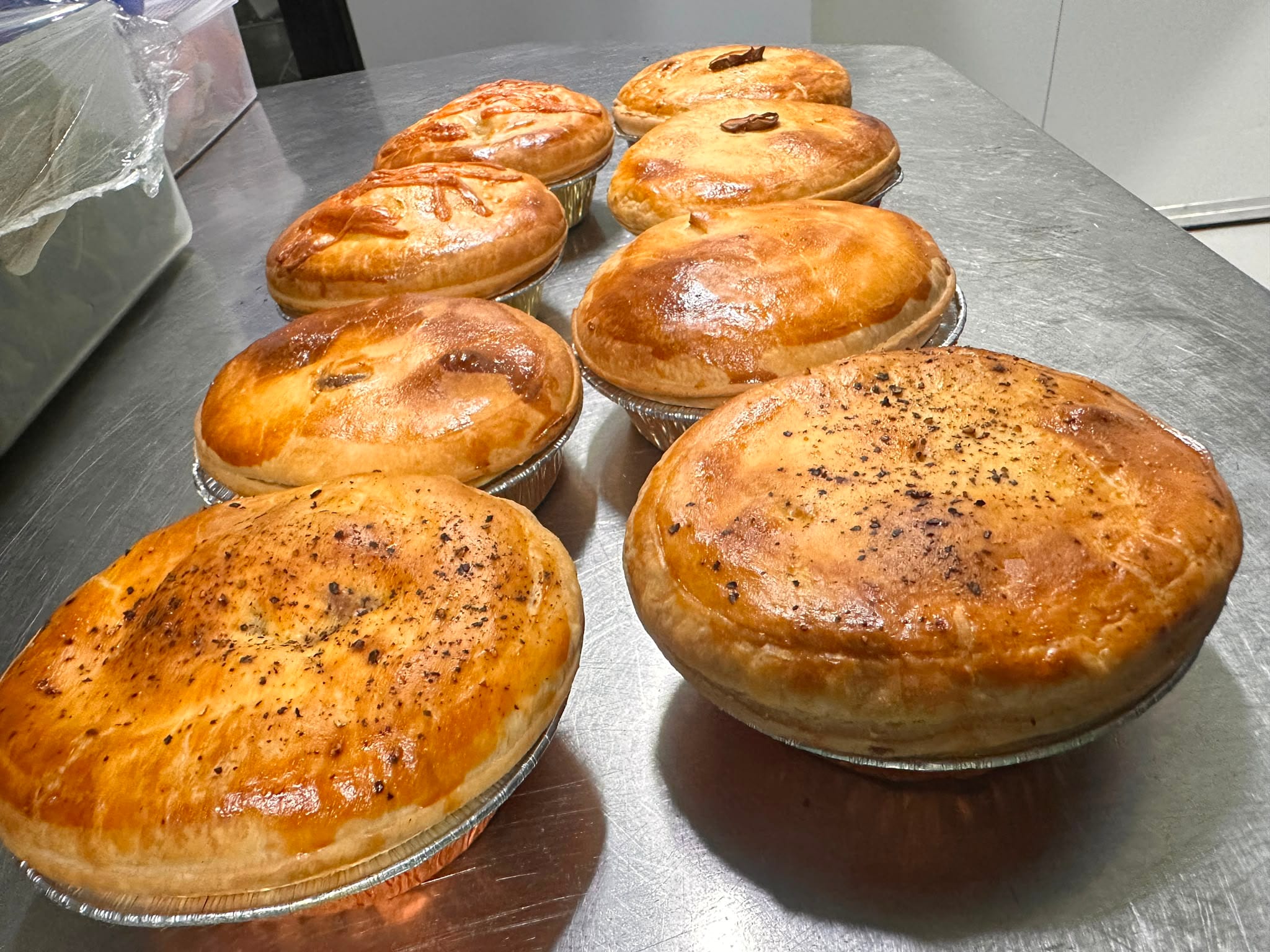 A row of pies in foil trays — pepper, chunky beef and chicken, glazed and cracked with black pepper